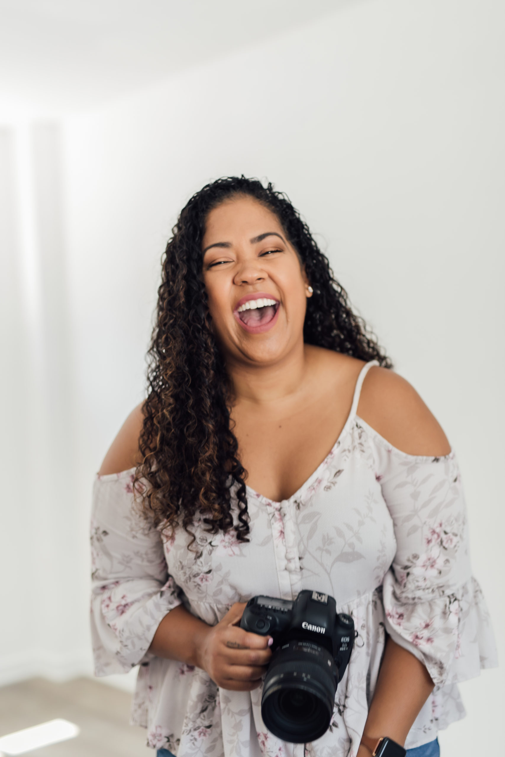 A joyful wedding photographer with curly hair holding a camera, laughing and wearing a floral print top in a bright white room.