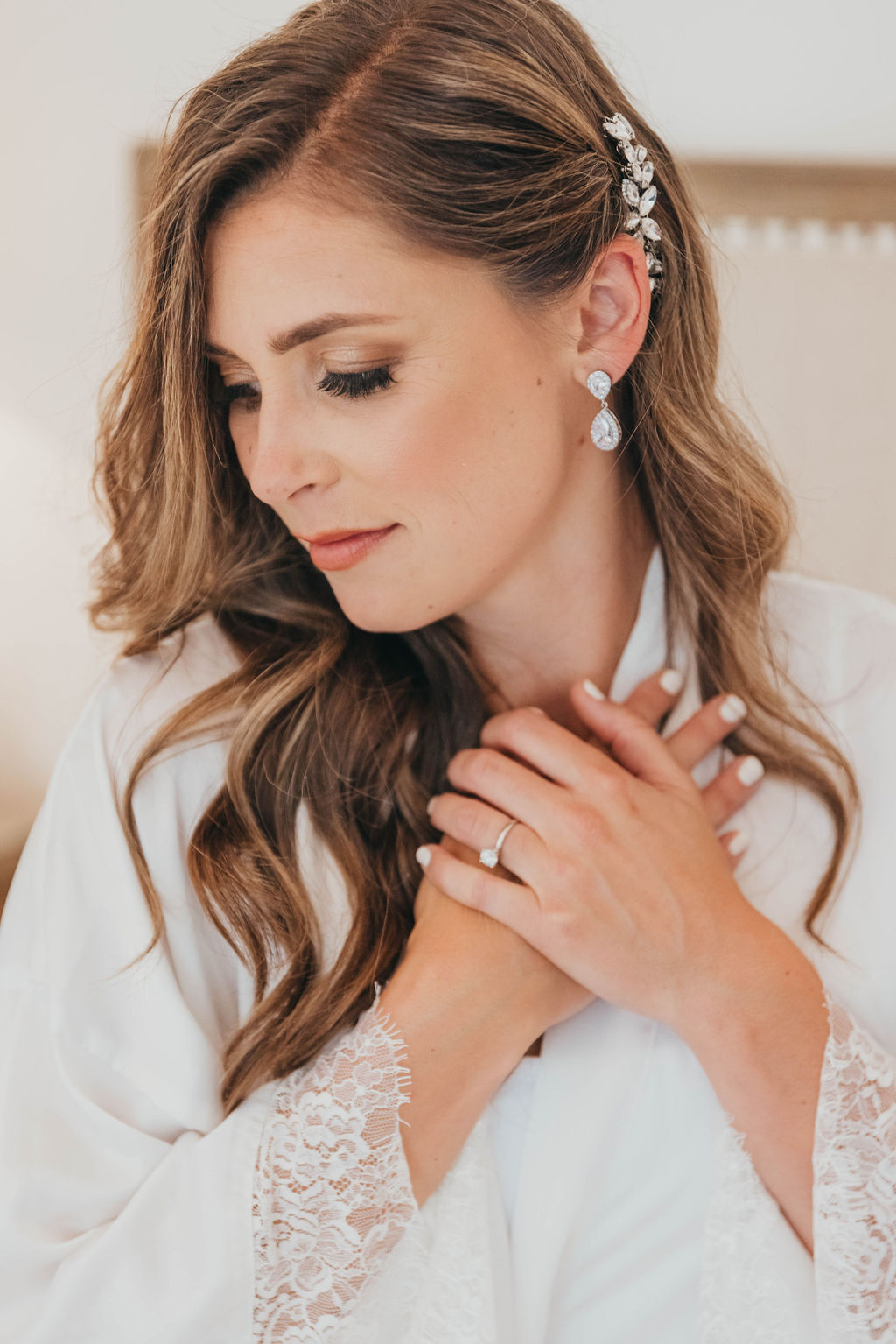 A bride in a white lace robe gently touches her collarbone, looking downwards, with a subtle smile, wearing a delicate hairpiece and earrings.
