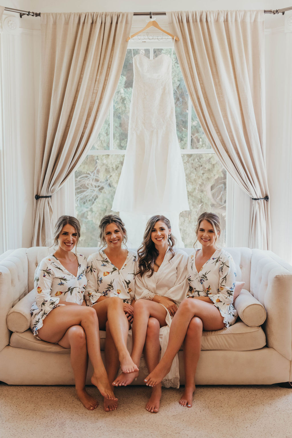 Bridesmaids in getting ready outfits matching floral robes laugh and converse on a white couch in a brightly lit room with large windows.