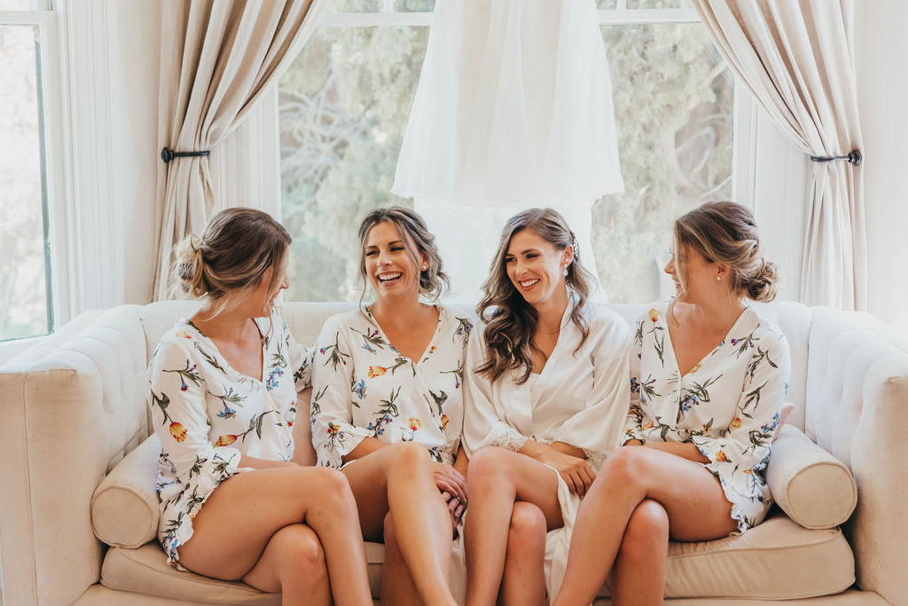 Bridesmaids in getting ready outfits matching floral robes laugh and converse on a white couch in a brightly lit room with large windows.