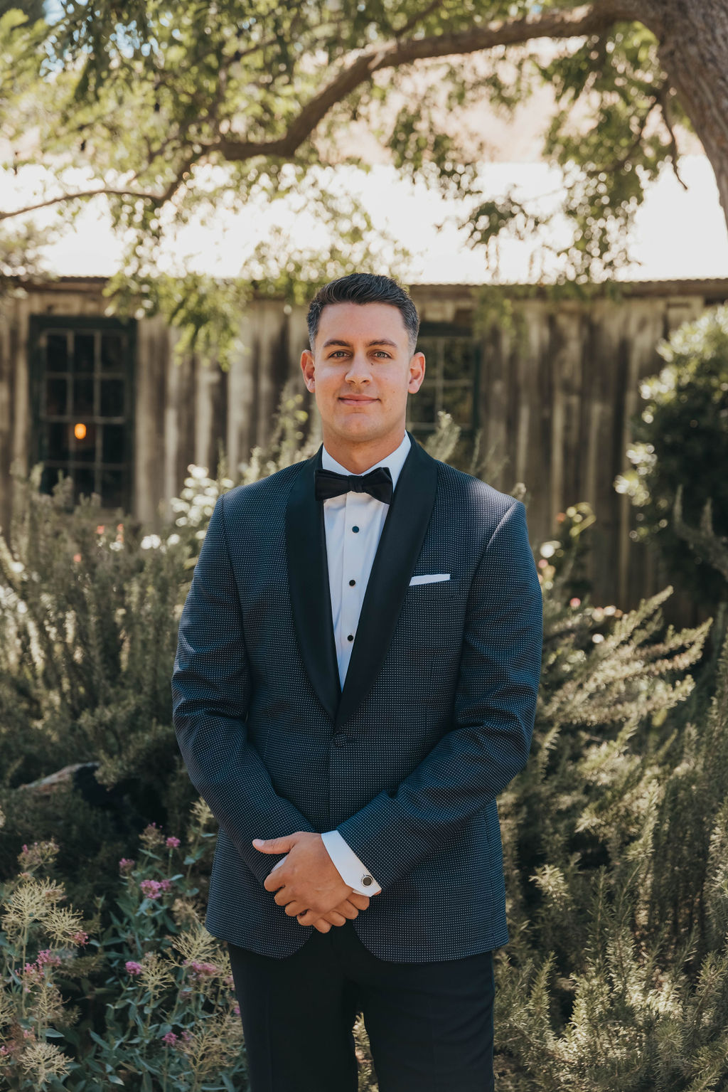 A man in a dark suit and bow tie standing confidently outdoors with a rustic wooden building and lush greenery in the background.