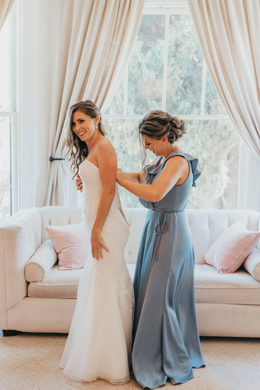 A woman in a blue dress adjusts the back of a bride's white gown in a bright room with large windows and elegant curtains at her Park Winters wedding
