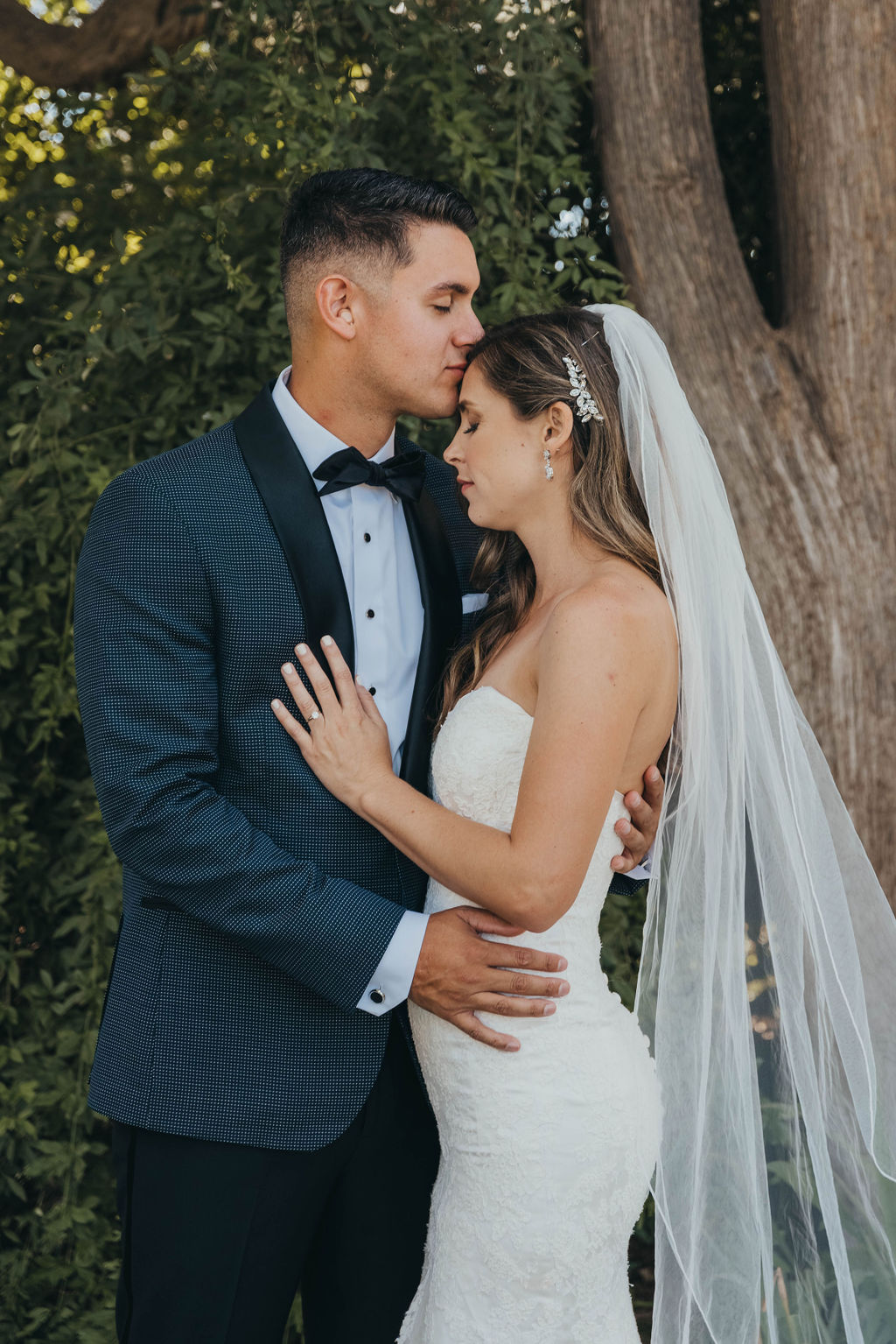 A bride in a white gown and a groom in a black tuxedo embrace, smiling, in a garden setting at their Park winters wedding
