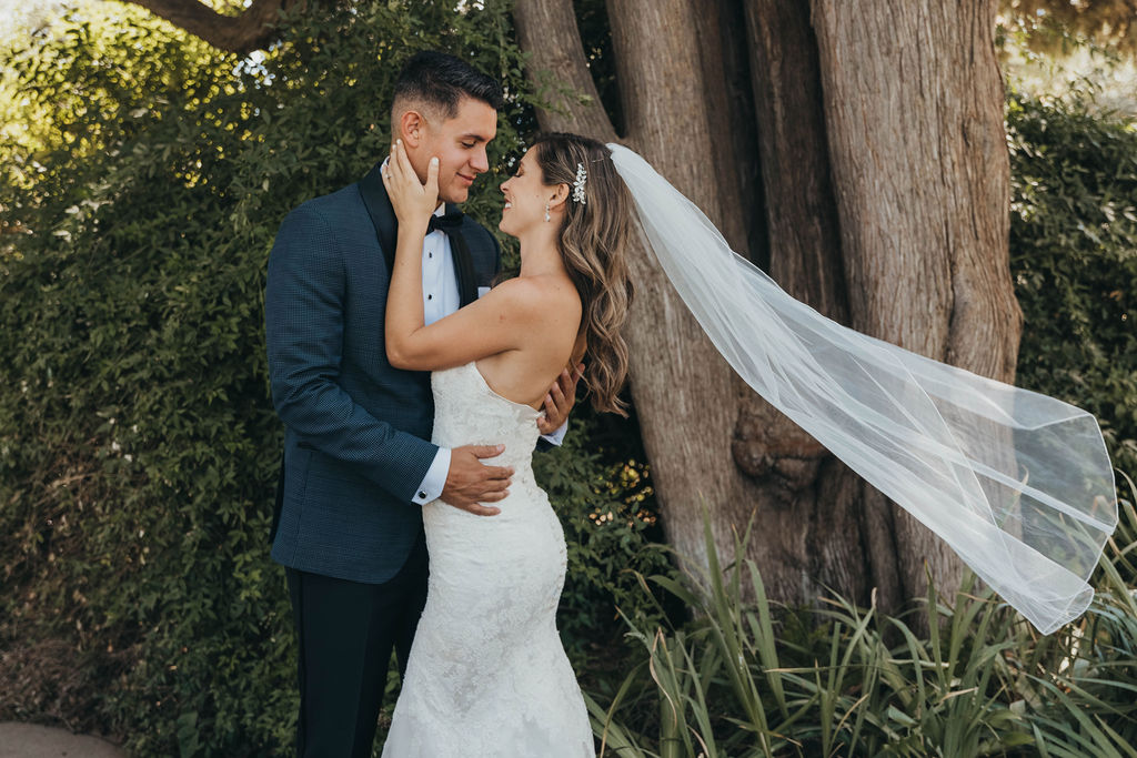 A bride in a white gown and a groom in a black tuxedo embrace, smiling, in a garden setting at their Park winters wedding