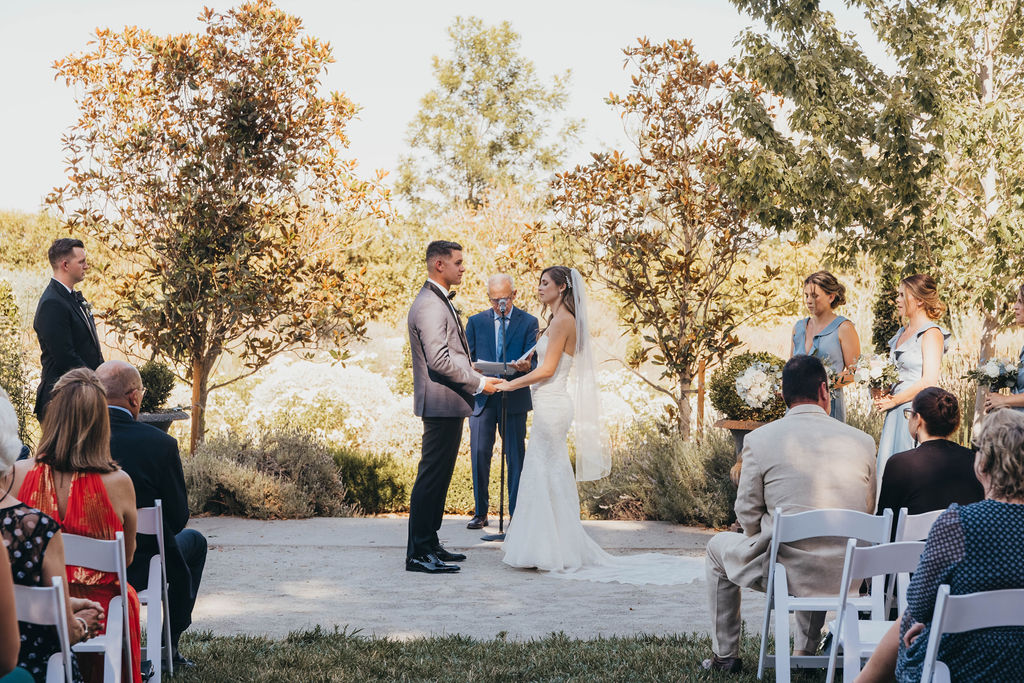 A park winters wedding ceremony outdoors with a couple exchanging vows in front of an officiant, surrounded by seated guests and trees.