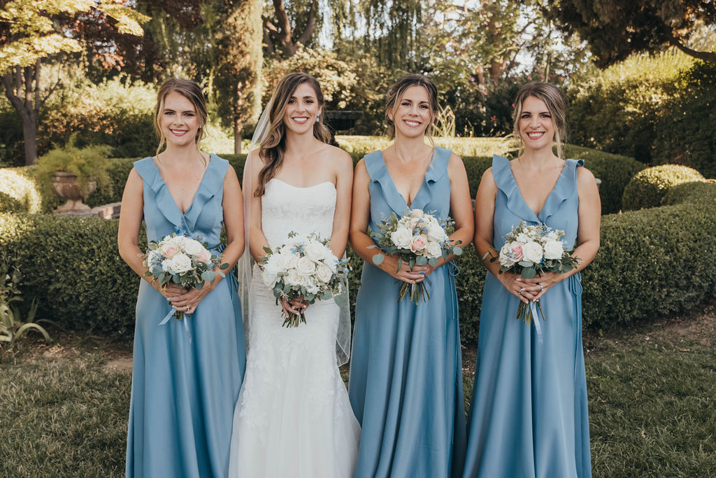 Four women dressed for a wedding, three in blue dresses and one in a white bridal gown, holding bouquets, standing in a garden.
