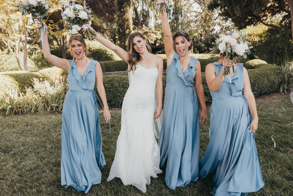 Four women dressed for a wedding, three in blue dresses and one in a white bridal gown, holding bouquets, standing in a garden.