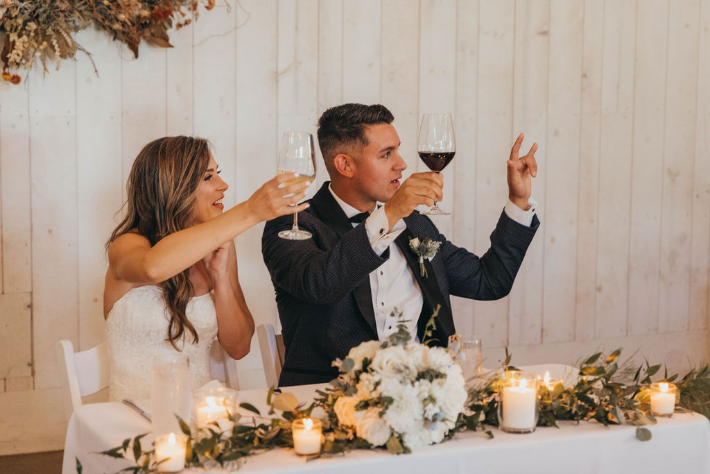 Bride and groom sitting at a table, toasting with wine glasses at a wedding reception, with candles and floral decorations around them.