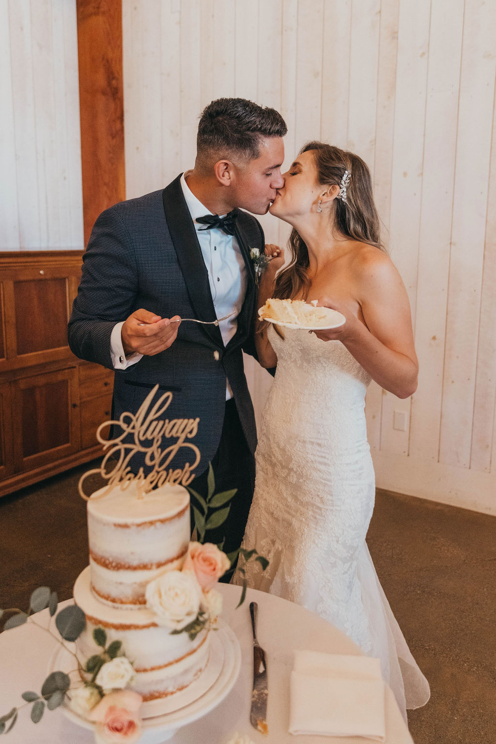 A newlywed couple kisses while holding a cake slice, standing beside a wedding cake with a topper reading "always together".