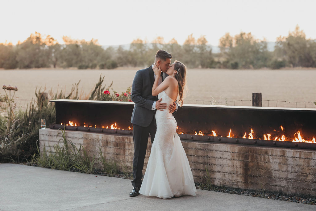 A bride and groom share a kiss along a fire pit outdoors at sunset.
