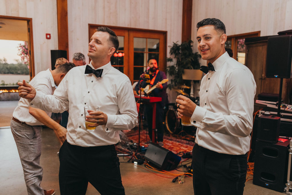 Two men in dress shirts and bow ties dancing at a lively indoor event, holding drinks, with a band playing in the background.