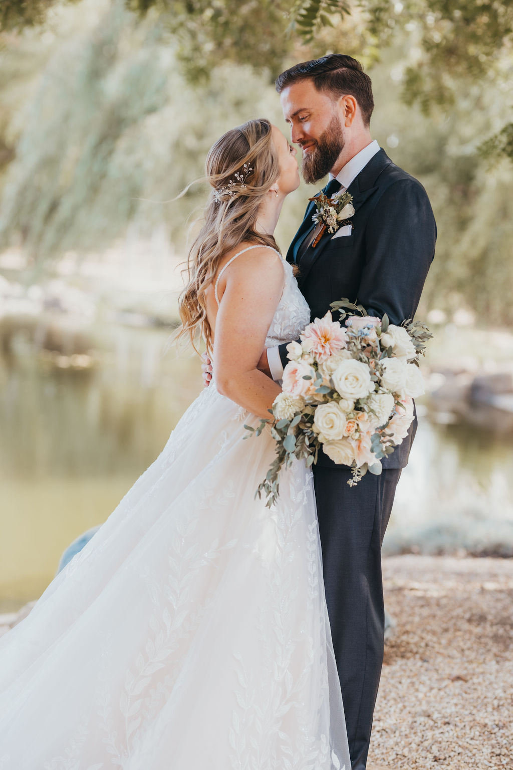 A bride and groom kissing beside a lake, surrounded by trees. the bride is holding a large bouquet of flowers.