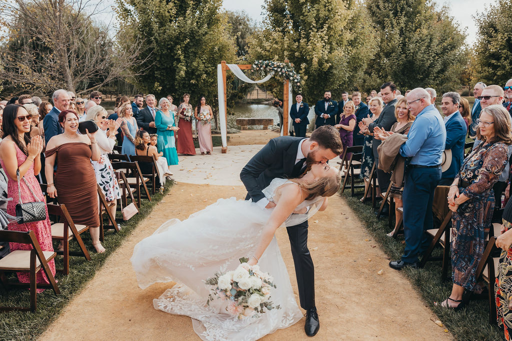 A bride and groom kissing beside a lake, surrounded by trees. the bride is holding a large bouquet of flowers. Captured by california wedding photographer