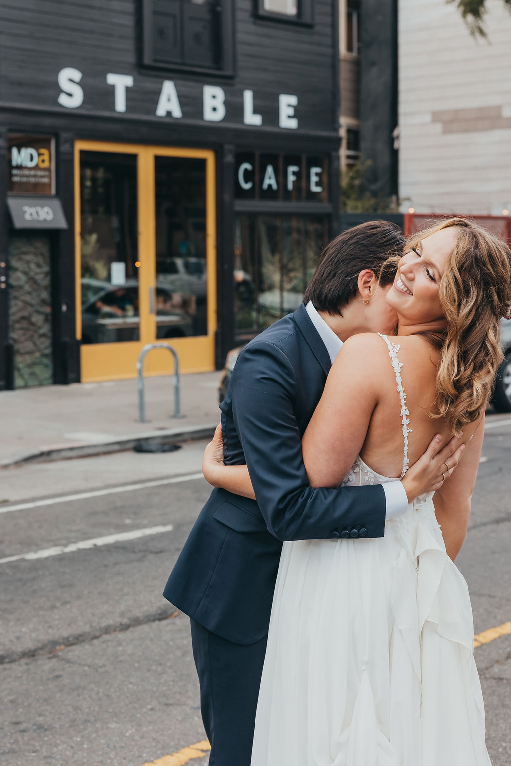 A couple in wedding attire embraces joyfully on a city street outside the stable cafe, 