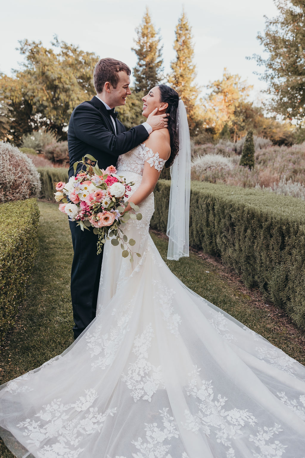 A bride and groom embrace in a garden, the bride holding a large bouquet and wearing a long lace dress, both smiling at each other.