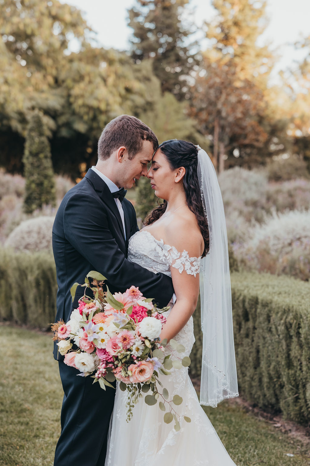 A bride and groom holding each other and looking into each other’s eyes, standing in a garden. the bride is holding a large bouquet of flowers.