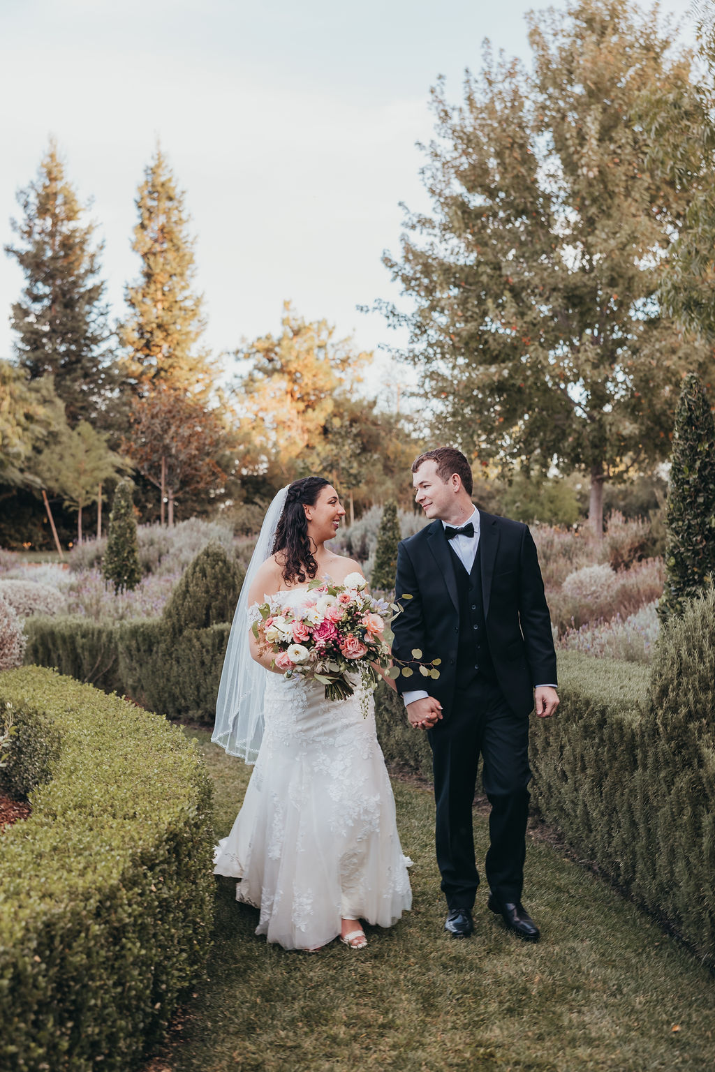 A bride and groom holding hands, walking down a tree-lined road, viewed from behind. the bride is wearing a long white gown and veil; the groom is in a black suit captured by california wedding photographer