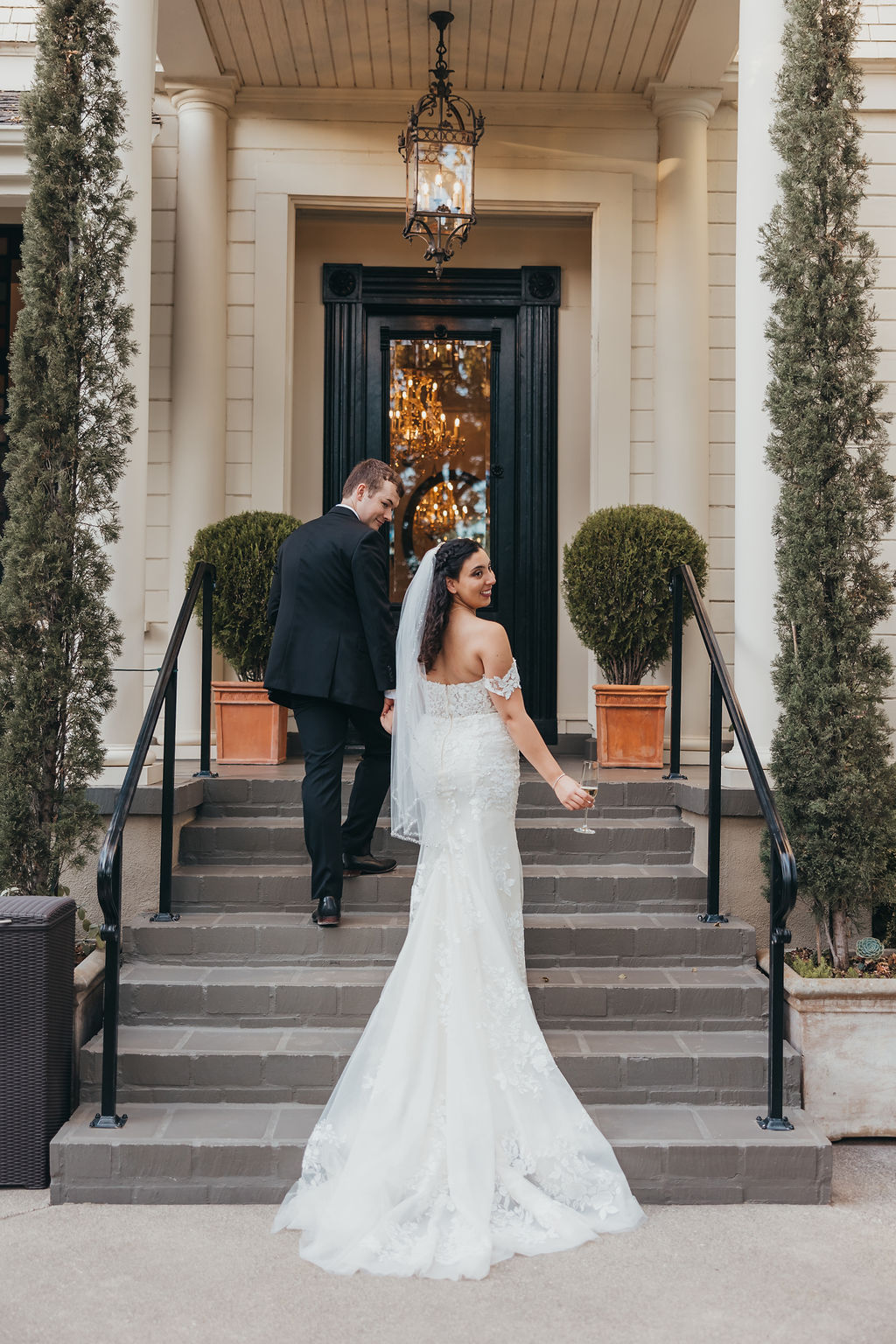 A bride and groom holding hands, walking down a tree-lined road, viewed from behind. the bride is wearing a long white gown and veil; the groom is in a black suit captured by california wedding photographer