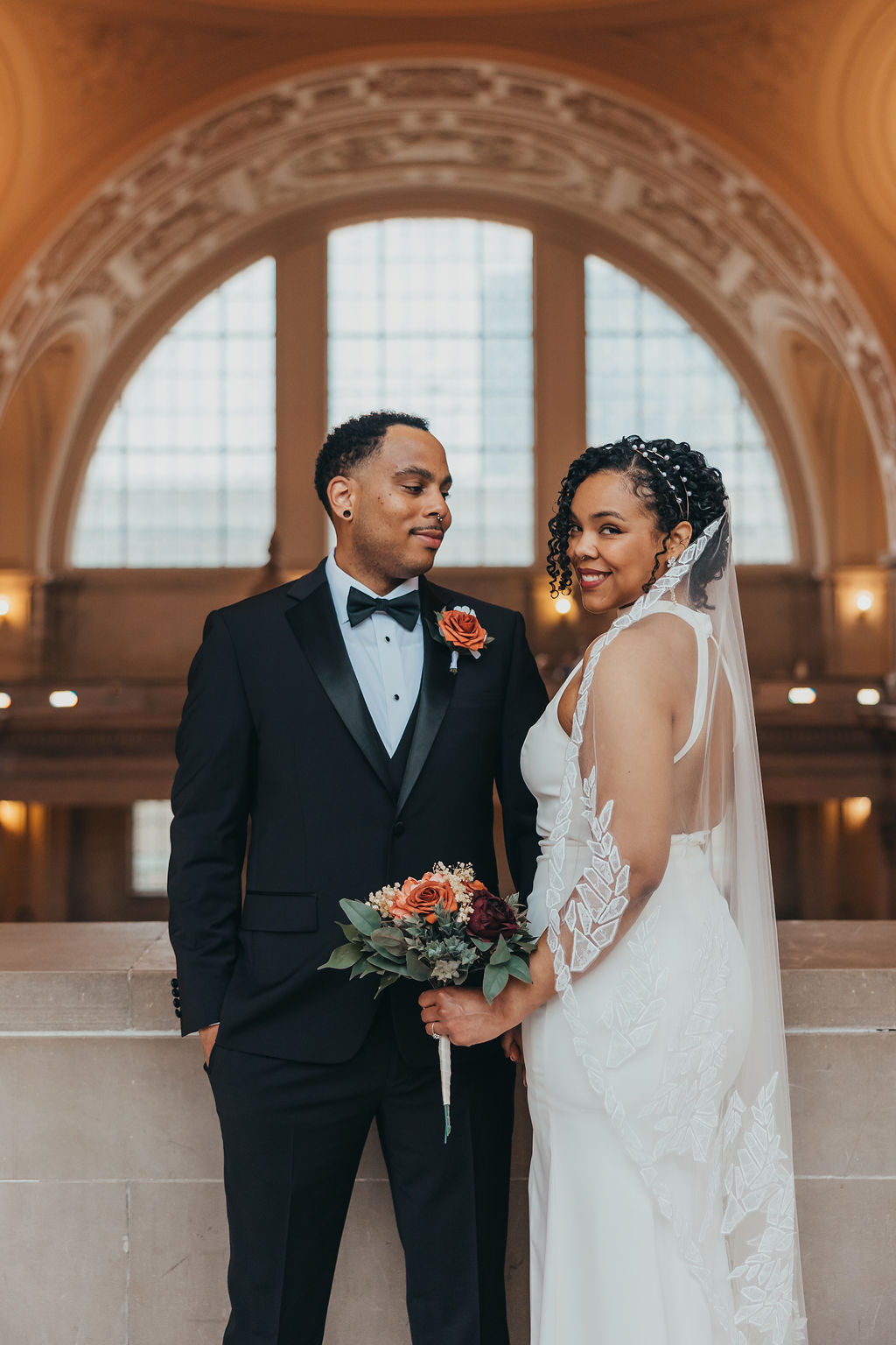 A bride and groom smiling at each other in a grand hall, the bride in a white dress and veil, the groom in a black tuxedo with a boutonniere at their wedding