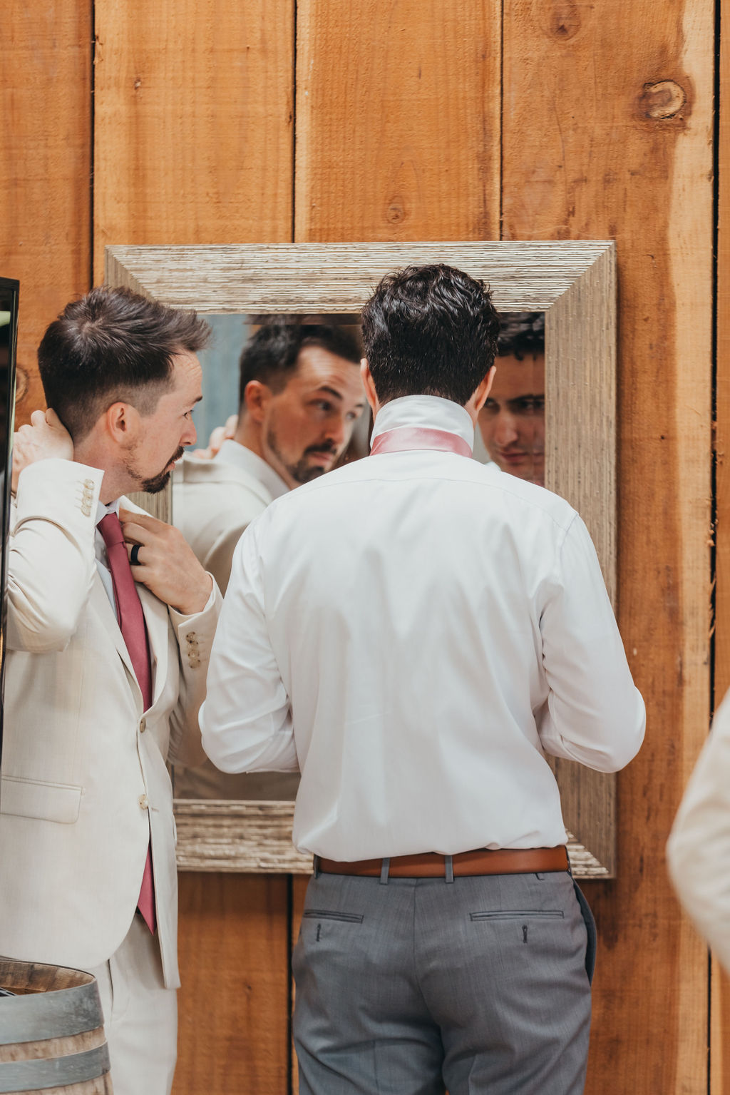 Two men in formal attire stand in front of a mirror. One adjusts his necktie while the other looks at his reflection. They are in a wooden-paneled room.