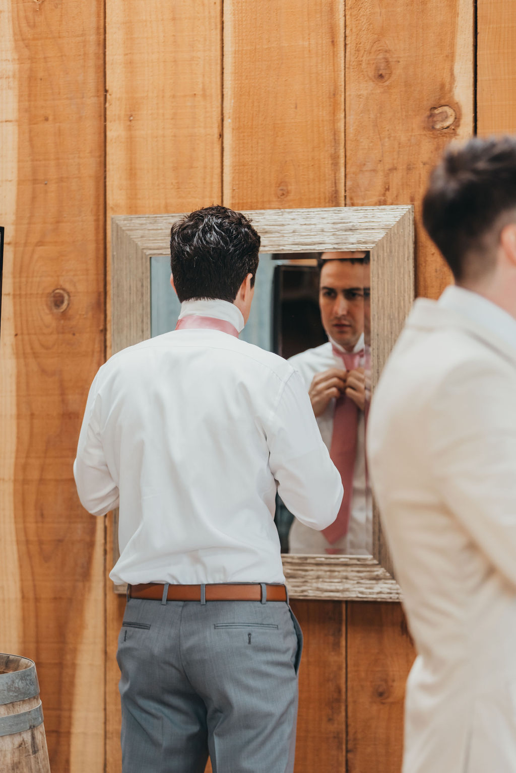 Two men in formal attire stand in front of a mirror. One adjusts his necktie while the other looks at his reflection. They are in a wooden-paneled room.