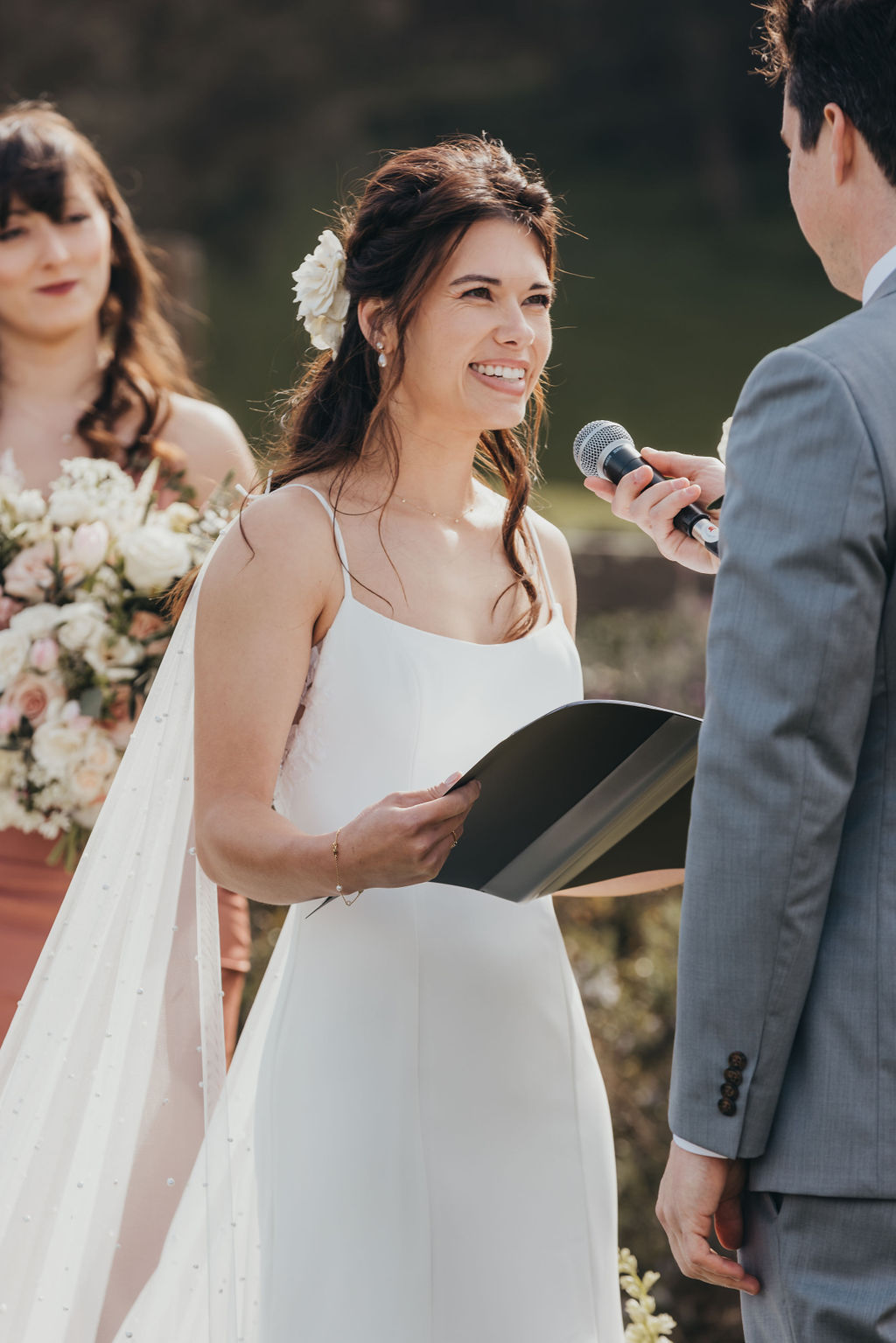 A bride and groom are standing outdoors, holding hands, and facing each other. The groom is wearing a light gray suit with a pink tie, and the bride is in a white dress with lace details and a flower in her hair at the haven at tomales