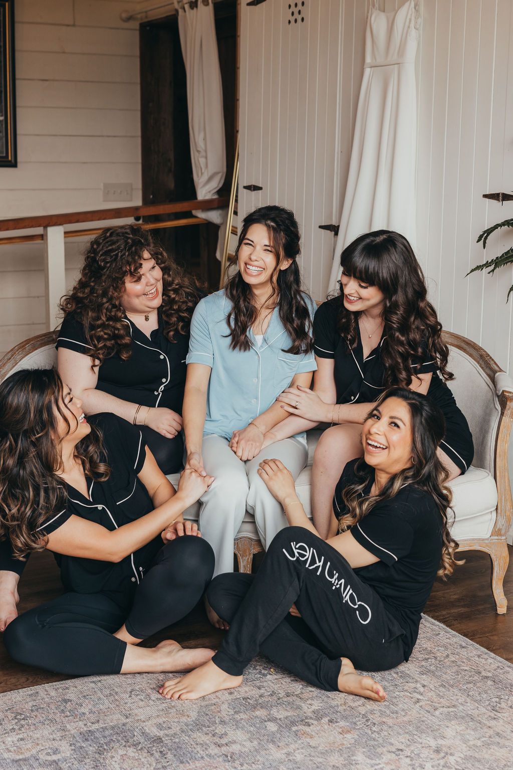 Five women sit and kneel in front of a white barn door, with a hanging wedding dress in the background. They are dressed in matching black pajamas, except one in blue. They are all smiling for a wedding