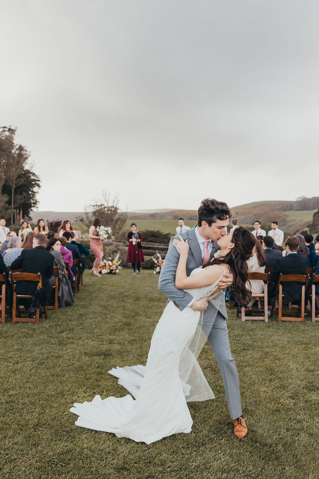 A bride and groom are standing outdoors, holding hands, and facing each other. The groom is wearing a light gray suit with a pink tie, and the bride is in a white dress with lace details and a flower in her hair at the haven at tomales