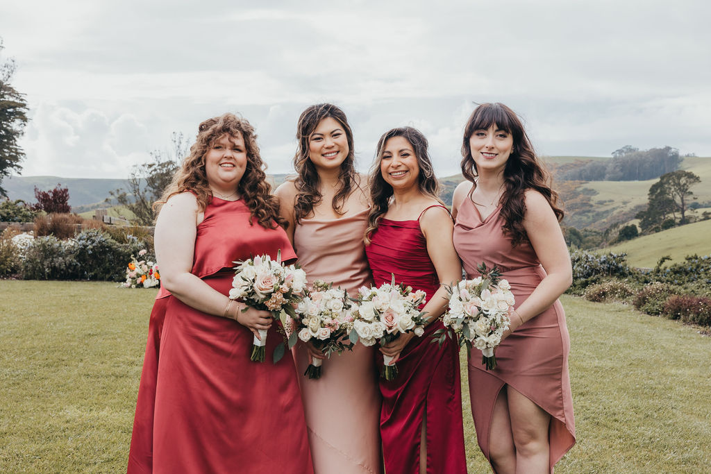 A bride in a white dress walks outdoors with four bridesmaids in long dresses of red and beige, holding bouquets, with a grassy landscape and cloudy sky in the background at the haven at tomales