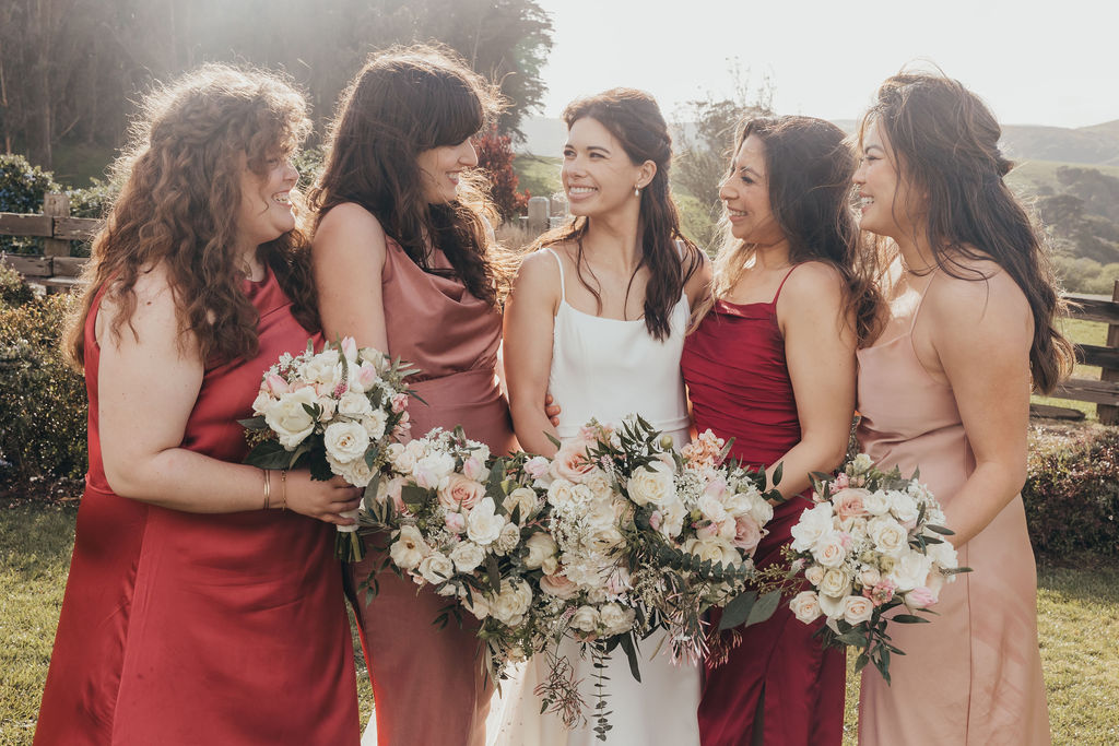 A bride in a white dress walks outdoors with four bridesmaids in long dresses of red and beige, holding bouquets, with a grassy landscape and cloudy sky in the background at the haven at tomales
