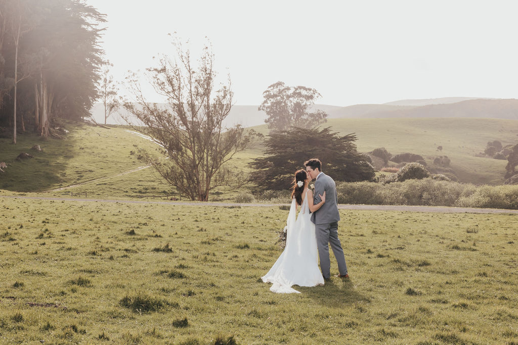 A bride and groom stand in a grassy field, with trees in the background and sunlight casting a soft glow over the scene at The Haven at Tomales