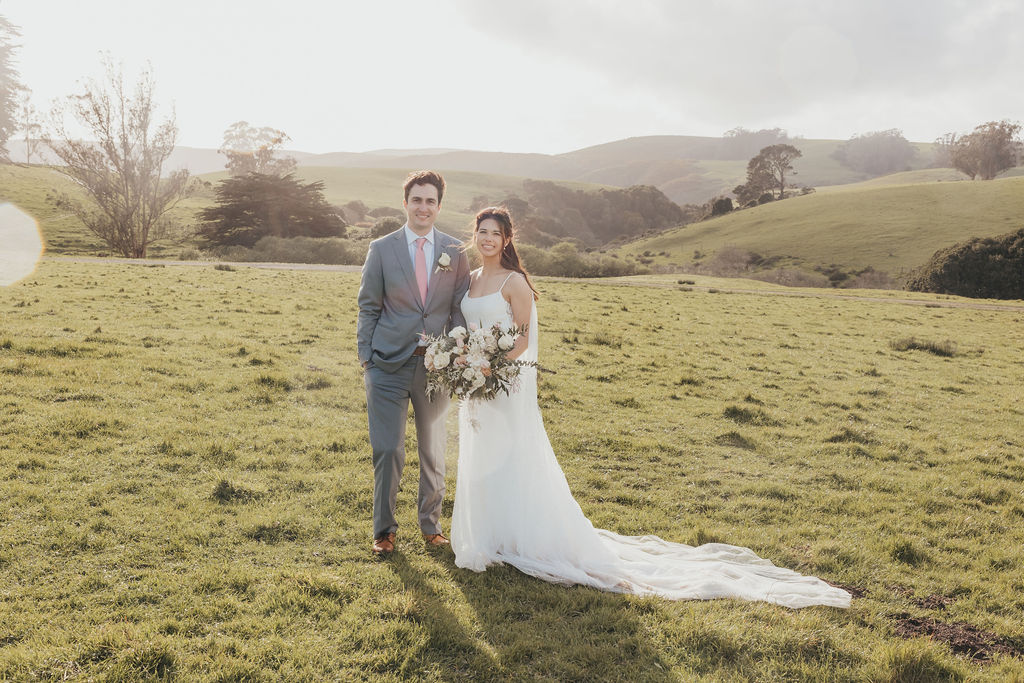 A bride and groom stand in a grassy field, with trees in the background and sunlight casting a soft glow over the scene at The Haven at Tomales