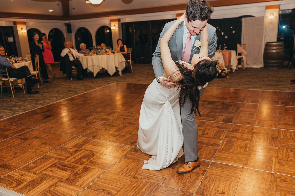 A bride and groom dance together on a parquet floor, surrounded by seated guests in a reception hall setting.