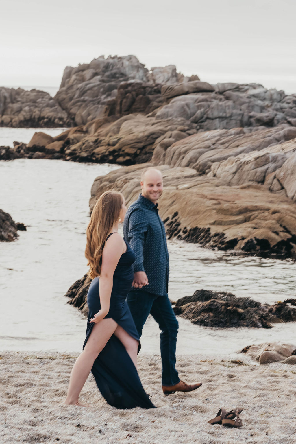 A couple stands close together embracing on a rocky beach with the ocean and large rocks in the background.