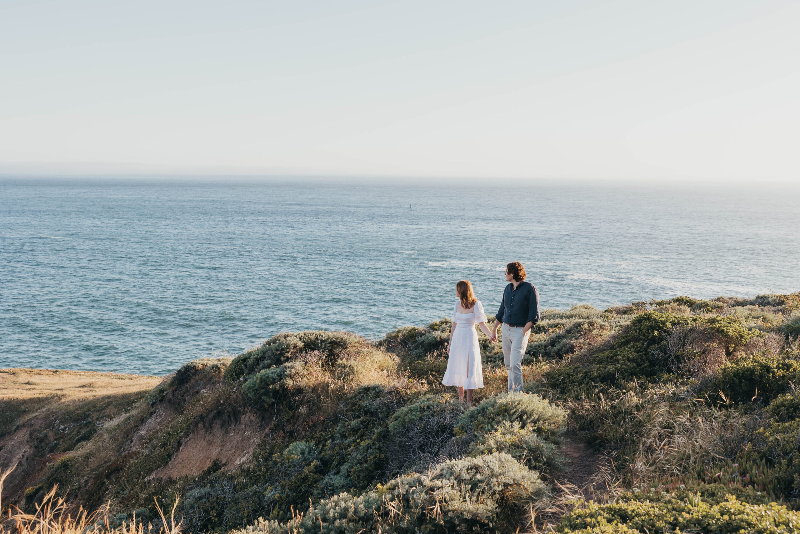 Two people stand on a grassy cliff overlooking the ocean on a clear day