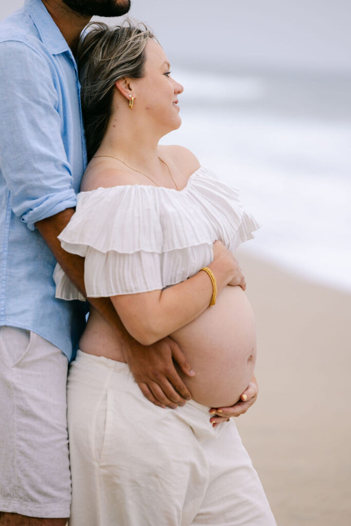 Expecting parents standing close together with ocean waves in the background at Half Moon Bay, California
